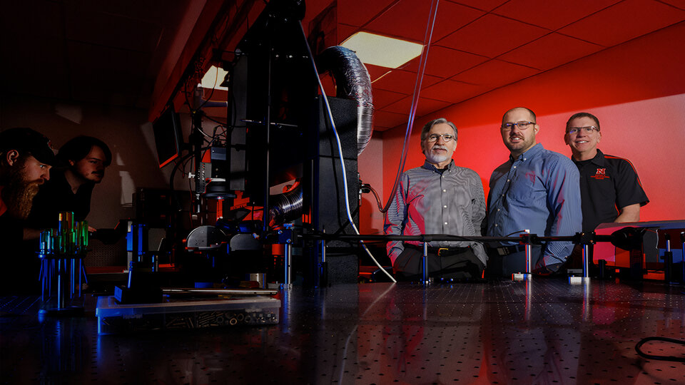Lab setting with equipment, glowing red light, and people standing under dim, dramatic lighting.