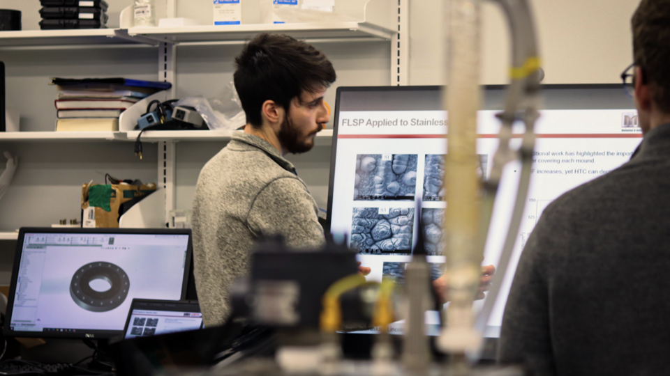 Person presenting research on a screen in a laboratory setting, with computers and equipment.